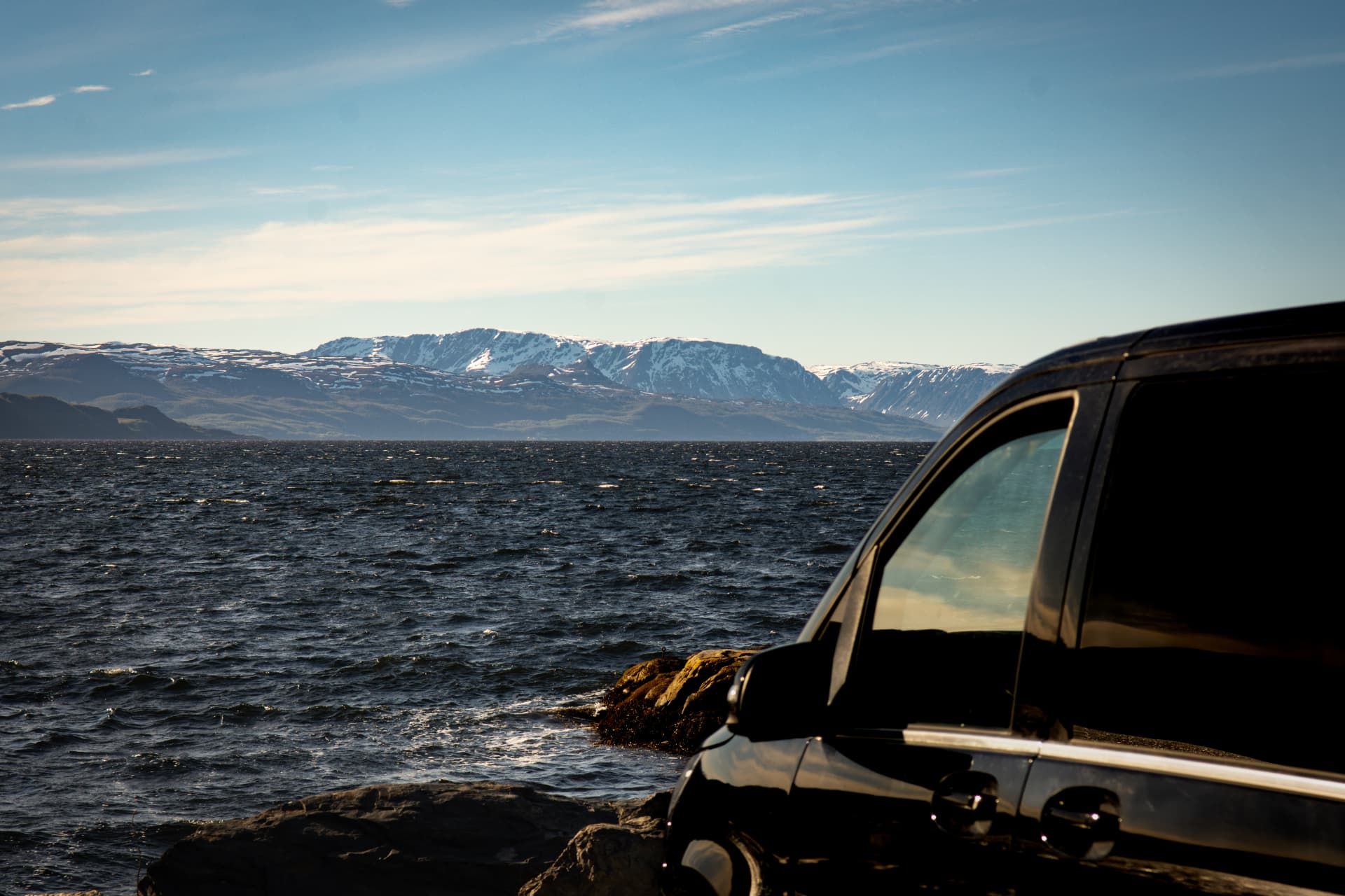 Premium black Mercedes V-Class parked on rock beside a fjord, snow-capped mountains across the water in Northern Norway