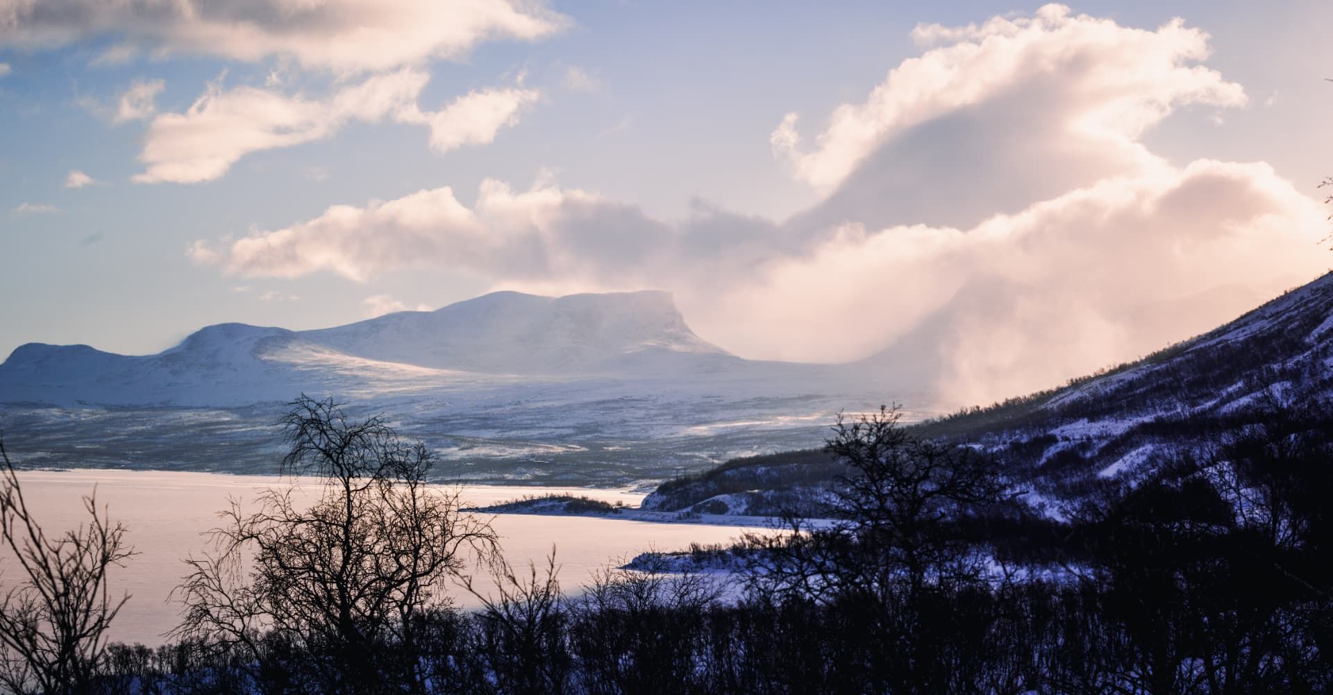 Wide winter view across a frozen lake towards distant mountains, with snow-covered slopes and soft clouds catching the light