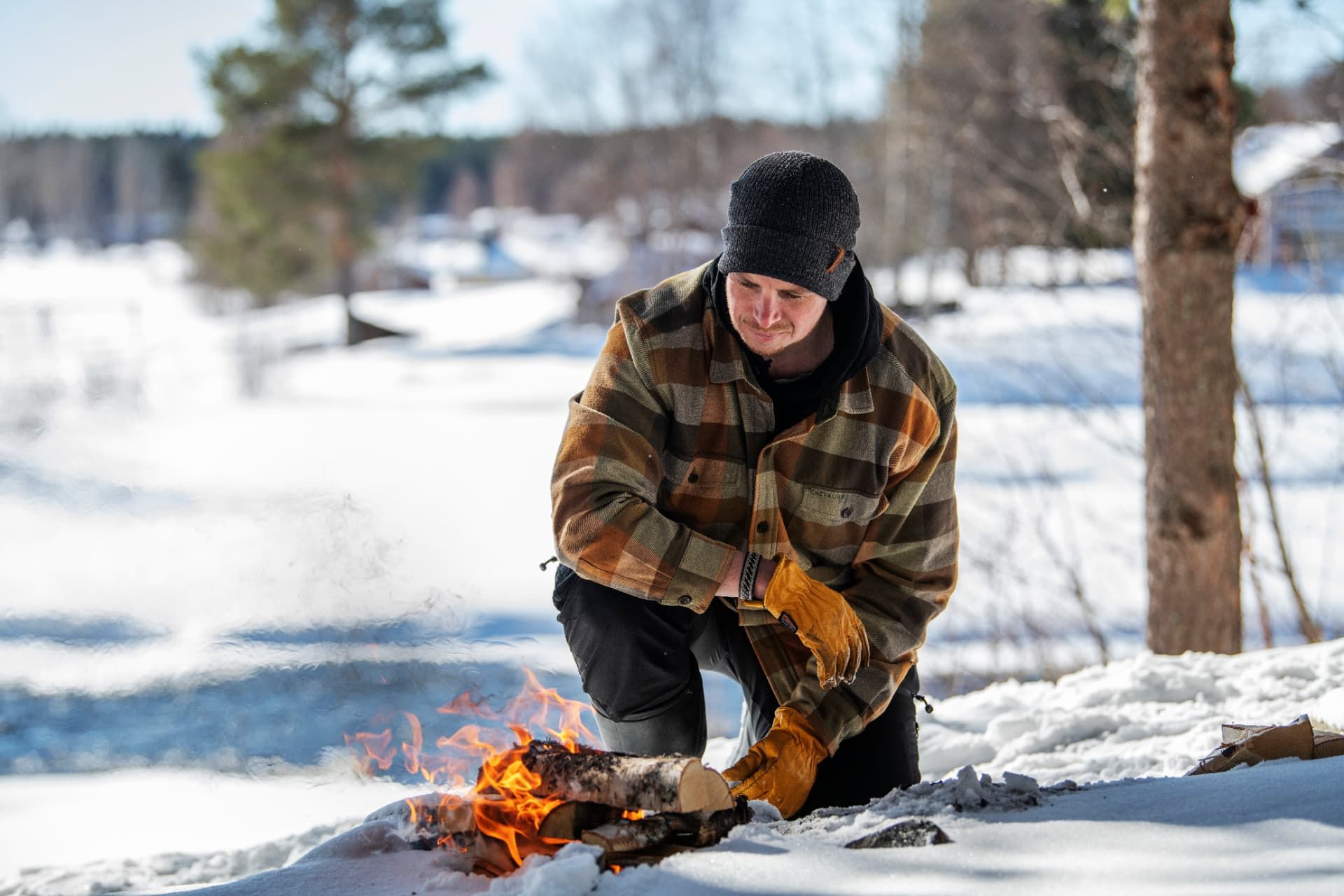 A man kneeling in the snow in front of a small campfire of logs, tending the fire during a lunch stop on a bright winter day